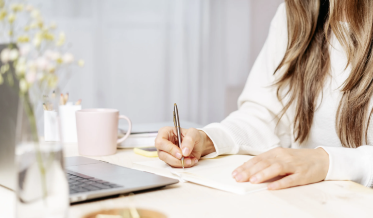 Person writing in a notebook beside a laptop and coffee mug on a bright workspace